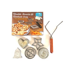 May include: A Double Rosette & Timbale Iron set including a brown cardboard box with product illustrations, a metal handle with a wooden grip, and five distinct metal molds in heart, star, bell, circular, and fluted round shapes.