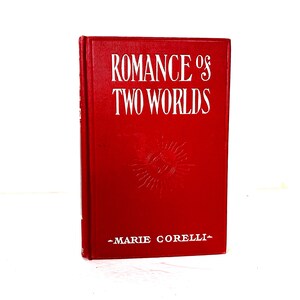 May include: A red hardcover book titled "ROMANCE of TWO WORLDS" with white lettering. The author's name, "MARIE CORELLI," is printed below the title. A sunburst design is in the center of the book.