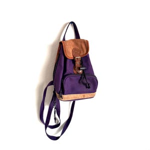 May include: A purple canvas backpack featuring a brown suede top flap and a matching suede base trim. The bag includes a front zippered pocket, a drawstring closure with a metal buckle, and adjustable purple shoulder straps for carrying.
