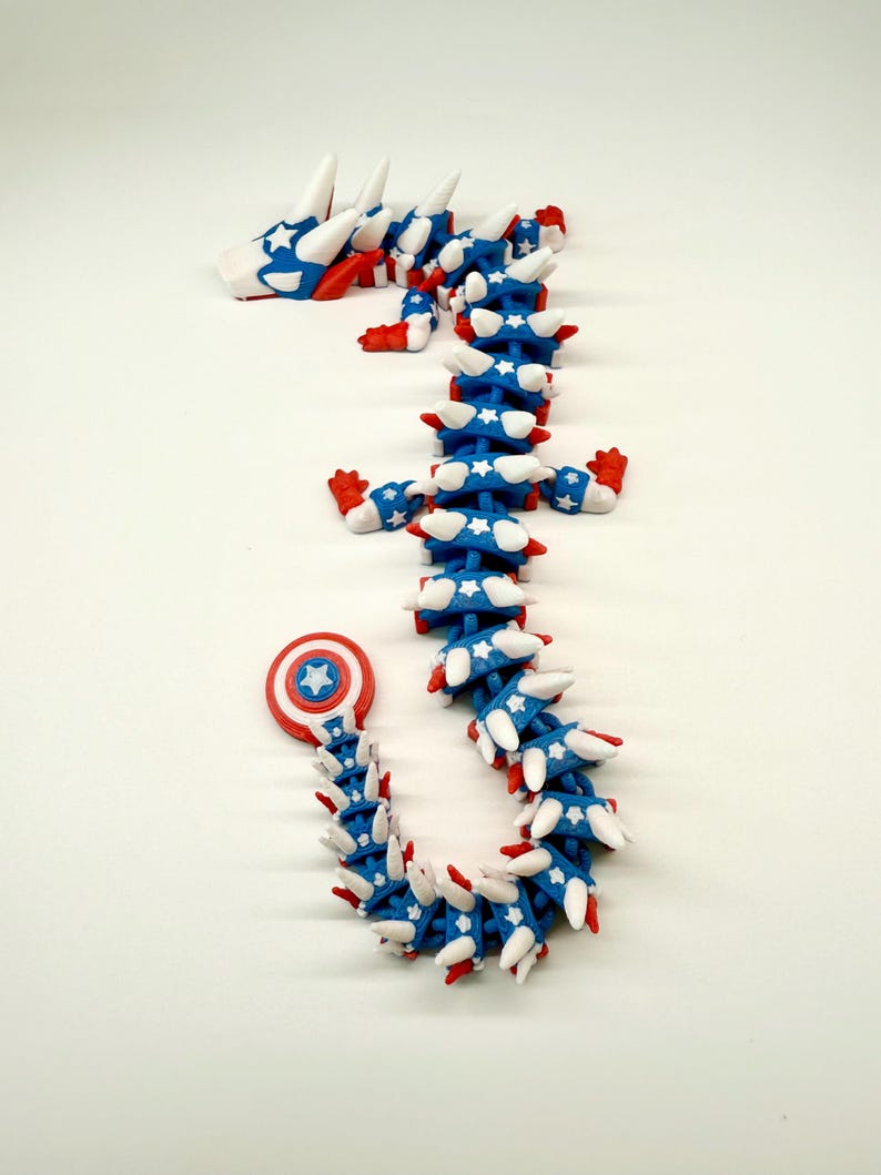 Puede incluir: Una escultura de drag&oacute;n articulada y caprichosa en colores patri&oacute;ticos. El drag&oacute;n presenta un cuerpo azul con estrellas blancas, detalles rojos y p&uacute;as blancas. La cabeza y la cola tambi&eacute;n est&aacute;n detalladas con la misma combinaci&oacute;n de colores. La cola del drag&oacute;n termina con un escudo.