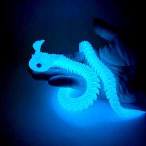 May include: A glowing blue articulated snake toy, held in a hand against a dark background. The toy has a detailed, textured surface and a stylized head with a single eye. The snake is curved and flexible, showcasing its segmented design.