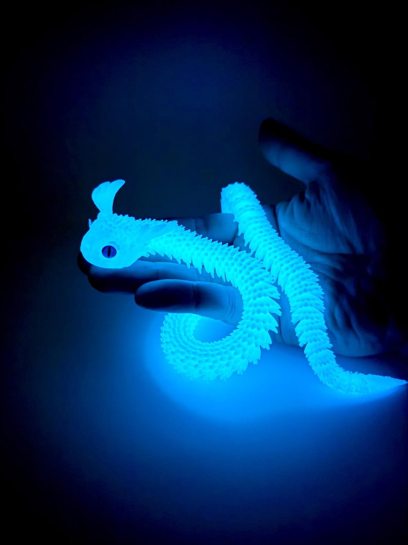 May include: A glowing blue articulated snake toy, held in a hand against a dark background. The toy has a detailed, textured surface and a stylized head with a single eye. The snake is curved and flexible, showcasing its segmented design.