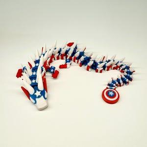 Puede incluir: Un juguete de drag&oacute;n articulado rojo, blanco y azul. El drag&oacute;n presenta un dise&ntilde;o patri&oacute;tico con estrellas y rayas. La cola termina con un escudo rojo, blanco y azul con una estrella. El drag&oacute;n mide aproximadamente 30 cm de largo.