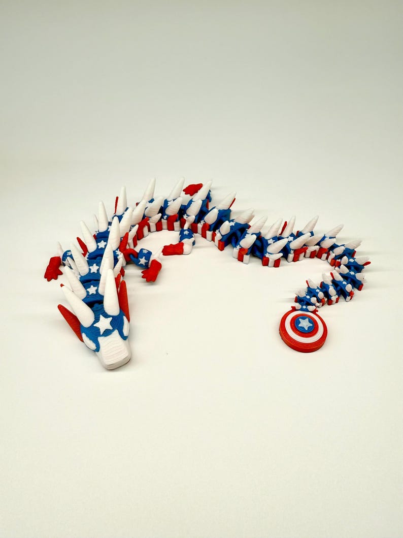 Puede incluir: Un juguete de drag&oacute;n articulado rojo, blanco y azul. El drag&oacute;n presenta un dise&ntilde;o patri&oacute;tico con estrellas y rayas. La cola termina con un escudo rojo, blanco y azul con una estrella. El drag&oacute;n mide aproximadamente 30 cm de largo.