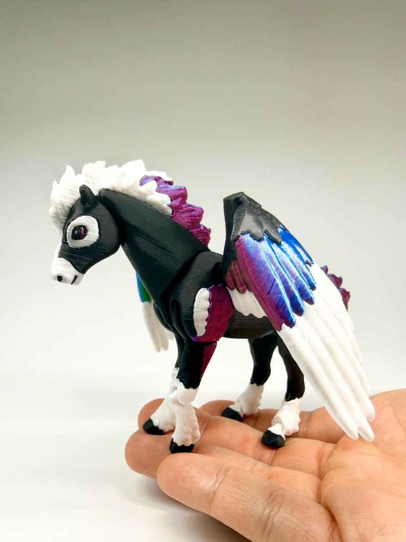 May include: A 3D-printed figurine of a pegasus, featuring a black body, white mane and tail, and wings with blue, purple, and white gradients. The horse has white legs with black hooves and is held in a hand.