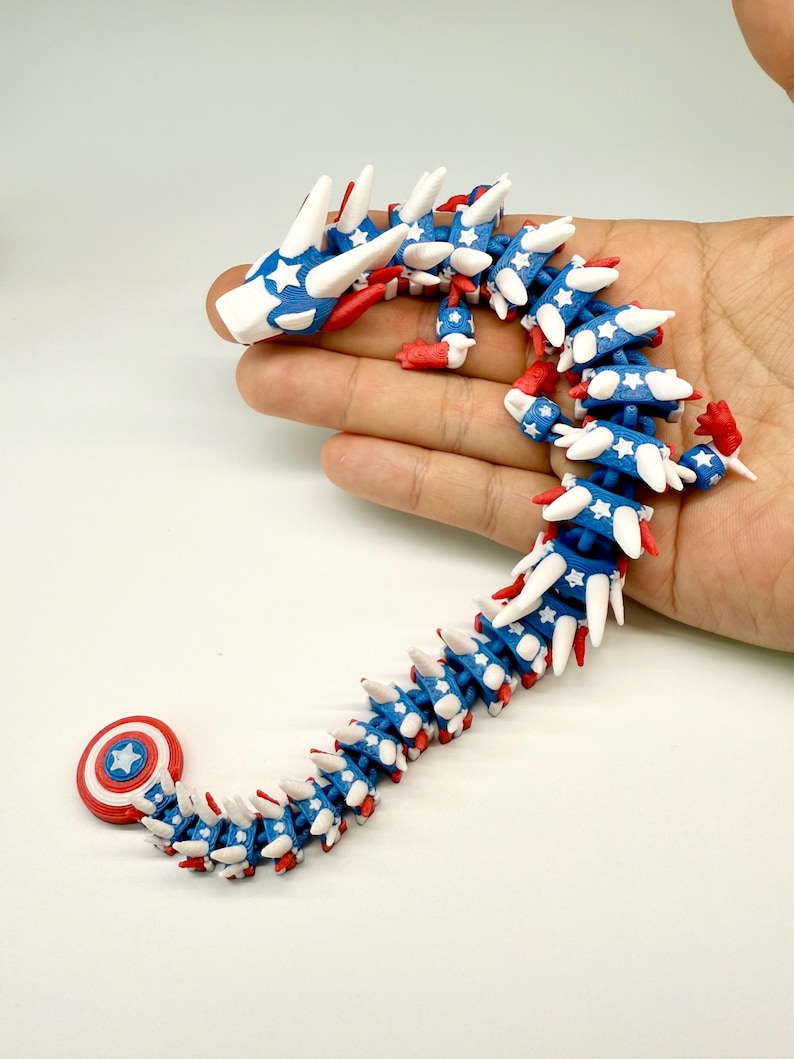 Puede incluir: Un juguete de drag&oacute;n articulado azul, rojo y blanco. El drag&oacute;n est&aacute; decorado con estrellas blancas sobre un fondo azul, con detalles rojos. La cabeza y la cola tambi&eacute;n son rojas y blancas. El drag&oacute;n se sostiene en una mano.