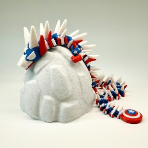 Puede incluir: Una figura de drag&oacute;n articulada impresa en 3D en rojo, blanco y azul, que se asemeja a la bandera estadounidense. El drag&oacute;n est&aacute; posado sobre una roca blanca texturizada y moteada. Un escudo del Capit&aacute;n Am&eacute;rica est&aacute; al final de la cola del drag&oacute;n.