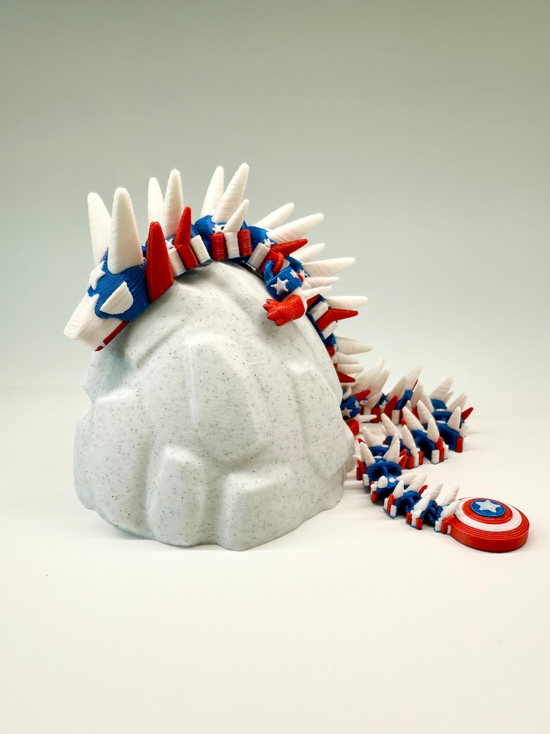 Puede incluir: Una figura de drag&oacute;n articulada impresa en 3D en rojo, blanco y azul, que se asemeja a la bandera estadounidense. El drag&oacute;n est&aacute; posado sobre una roca blanca texturizada y moteada. Un escudo del Capit&aacute;n Am&eacute;rica est&aacute; al final de la cola del drag&oacute;n.