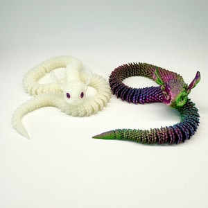 May include: Two articulated dragon toys. One is white with pink eyes, the other is a rainbow of colors including green, purple, and blue. Both have a textured, scale-like appearance and are posed on a white surface.