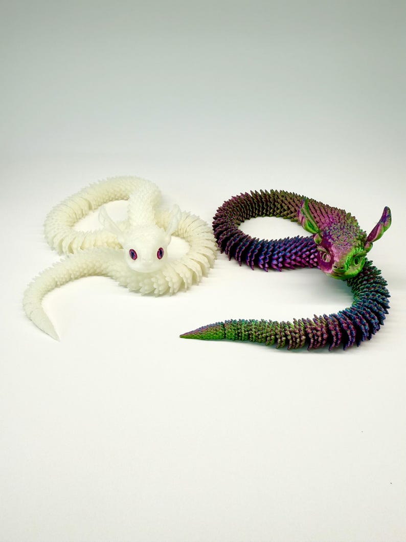 May include: Two articulated dragon toys. One is white with pink eyes, the other is a rainbow of colors including green, purple, and blue. Both have a textured, scale-like appearance and are posed on a white surface.