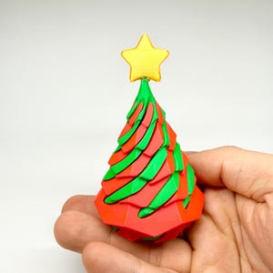 May include: A small, colorful Christmas tree decoration. The tree is red and green with a yellow star on top. The tree is held in a person's hand.