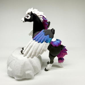 May include: A black and white pegasus figurine with white wings and a multi-colored mane and tail. The wings are white with blue and purple accents. The figurine is perched on a textured, light gray rock.