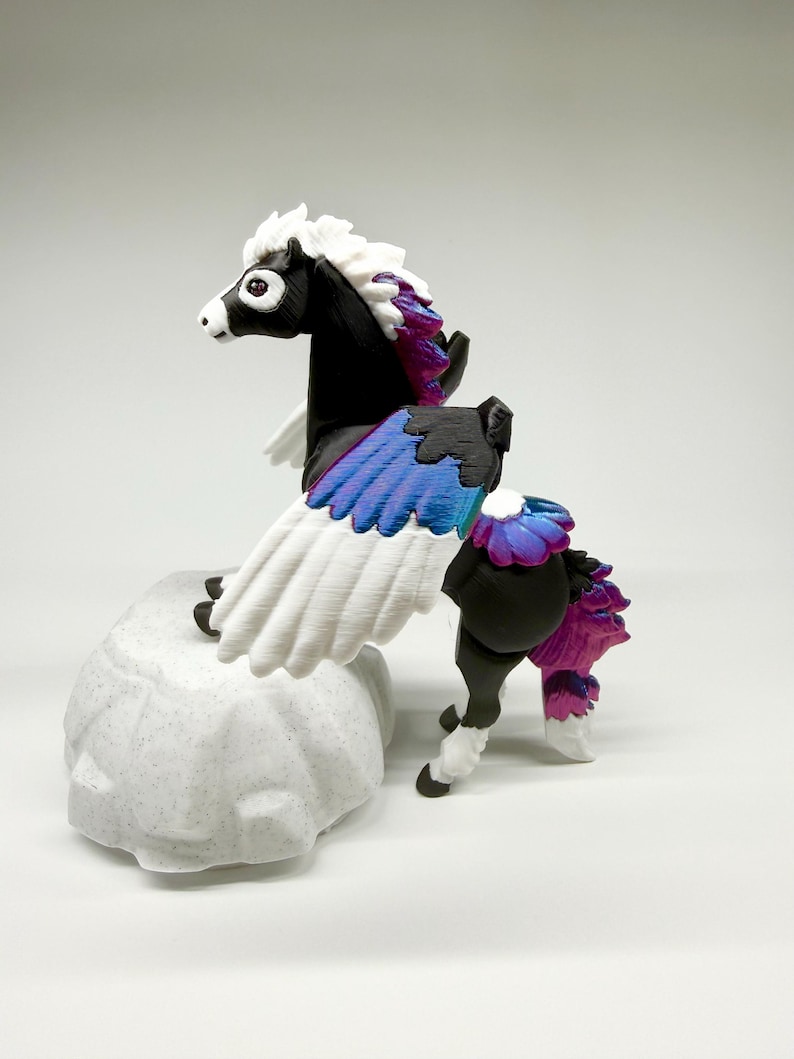 May include: A black and white pegasus figurine with white wings and a multi-colored mane and tail. The wings are white with blue and purple accents. The figurine is perched on a textured, light gray rock.