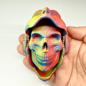Peut inclure: Une sculpture de crâne imprimée en 3D, multicolore, ornée d'un chapeau et d'un casque. Le crâne présente un mélange de couleurs arc-en-ciel avec des détails de lignes complexes. La sculpture est tenue dans une main, mettant en valeur sa taille et son design.