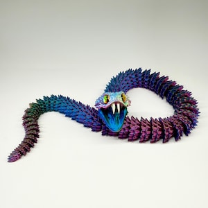 May include: A colorful, articulated snake toy in shades of blue, purple, and green. The snake's head is open, revealing white fangs and yellow eyes. The body is segmented, creating a flexible, textured appearance. The toy is set against a white background.