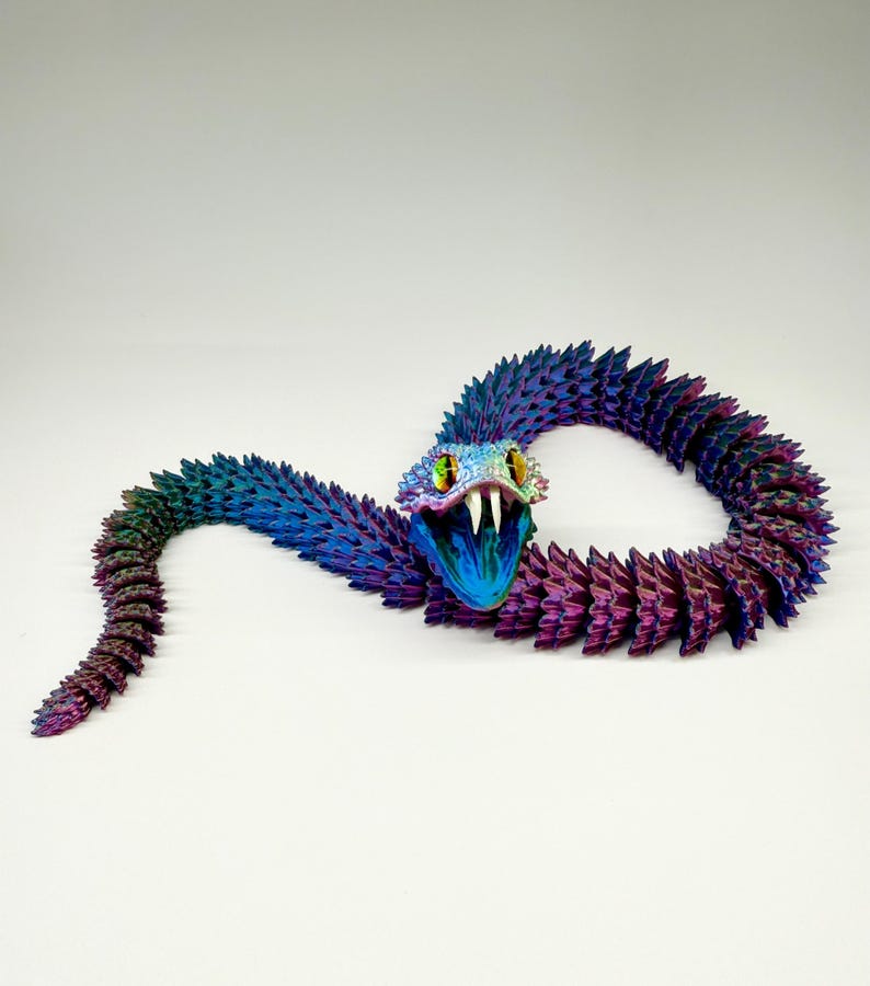 May include: A colorful, articulated snake toy in shades of blue, purple, and green. The snake's head is open, revealing white fangs and yellow eyes. The body is segmented, creating a flexible, textured appearance. The toy is set against a white background.