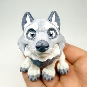Pode incluir: Figura detalhada, impressa em 3D, de um cão husky. O cão tem uma pelagem cinza e branca, com detalhes em cinza escuro nas orelhas e no nariz. A figura é segurada em uma mão, mostrando os detalhes intrincados do design.