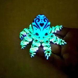 May include: A glowing octopus figurine in blue and green. The head features a detailed design, with heart-shaped patterns. The tentacles have black spots. The figurine is illuminated, creating a vibrant effect. The overall design is stylized and eye-catching.