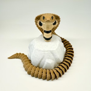 Op de afbeelding: Een gedetailleerde sculptuur van een cobra-slang in tinten bruin, zwart en wit. De kop van de slang is omhoog, met zijn kap uitgevouwen, en hij rust op een gespikkelde witte rots. Het lichaam van de slang krult rond de rots.