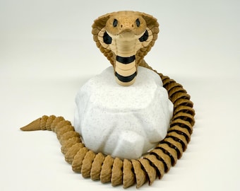 May include: A detailed sculpture of a cobra snake in shades of tan, black, and white. The snake's head is raised, with its hood expanded, and it rests on a speckled white rock. The snake's body curves around the rock.