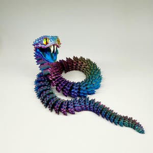 May include: A colorful, articulated snake toy in shades of blue, purple, and green. The snake has a detailed head with yellow eyes and white fangs. The body is segmented, allowing for flexible movement. The toy is posed on a white surface.