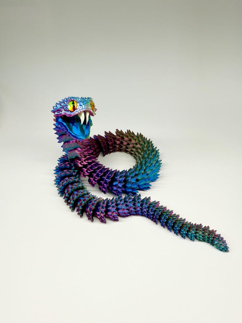 May include: A colorful, articulated snake toy in shades of blue, purple, and green. The snake has a detailed head with yellow eyes and white fangs. The body is segmented, allowing for flexible movement. The toy is posed on a white surface.