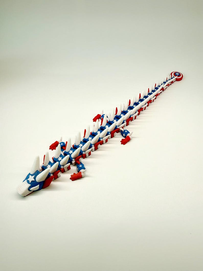 Puede incluir: Un juguete de drag&oacute;n articulado rojo, blanco y azul. El drag&oacute;n presenta una cabeza y un cuerpo con patrones de estrellas, detalles en rojo y blanco y una cola circular. El juguete es de pl&aacute;stico y es posable.