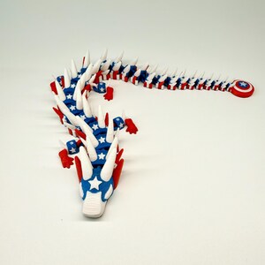 Puede incluir: Un juguete de drag&oacute;n articulado rojo, blanco y azul. El drag&oacute;n tiene un cuerpo segmentado, con un patr&oacute;n de estrellas azul y blanco en la cabeza y el cuerpo. Las p&uacute;as blancas recorren la columna vertebral y un escudo rojo con una estrella est&aacute; en la cola.