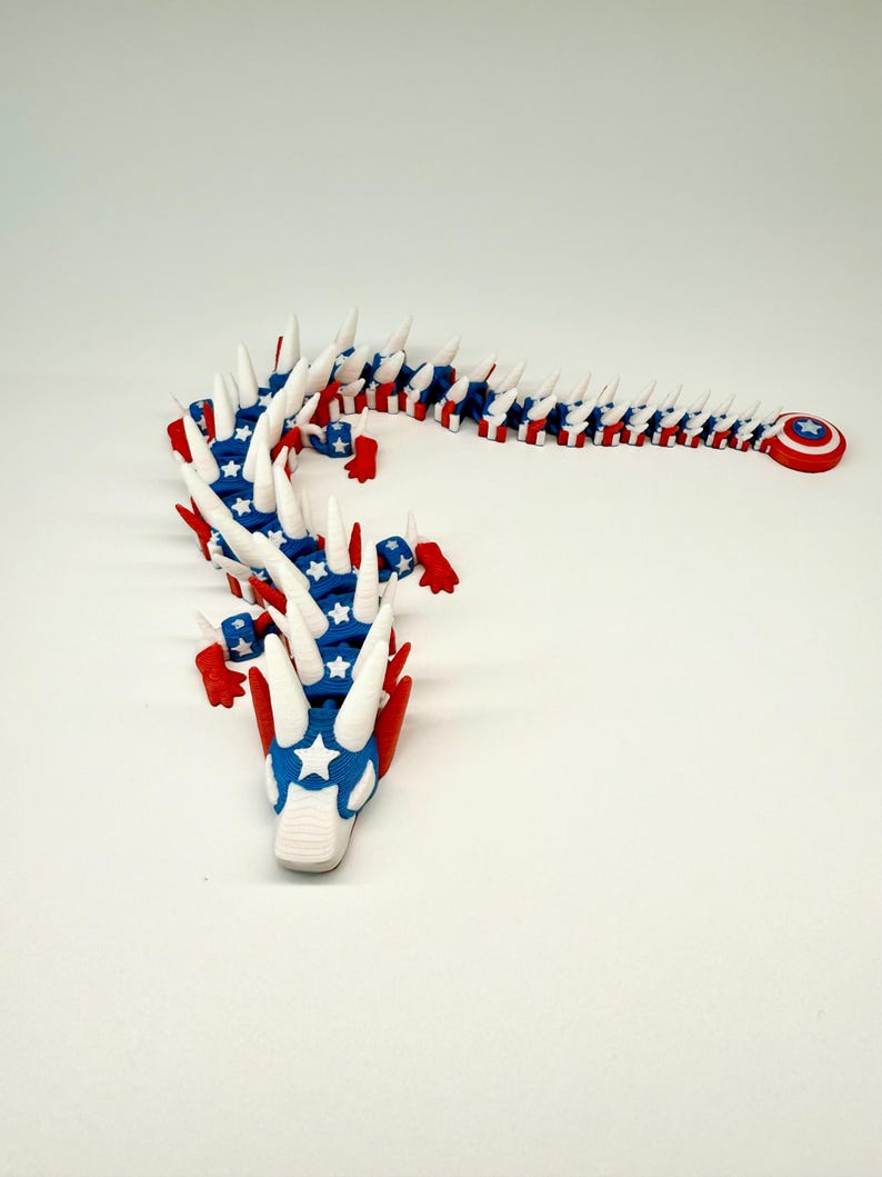 Puede incluir: Un juguete de drag&oacute;n articulado rojo, blanco y azul. El drag&oacute;n tiene un cuerpo segmentado, con un patr&oacute;n de estrellas azul y blanco en la cabeza y el cuerpo. Las p&uacute;as blancas recorren la columna vertebral y un escudo rojo con una estrella est&aacute; en la cola.