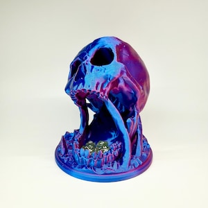 Op de afbeelding: Een 3D-geprinte dobbelsteenhouder in de vorm van een schedel in blauwe en paarse tinten. De schedel heeft open oogkassen en een brede mond, met dobbelstenen erin. De basis is een getextureerd, cirkelvormig platform.