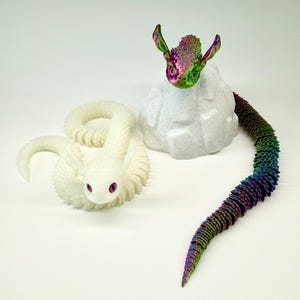 May include: Two fantasy dragon sculptures. One is white with pink eyes, coiled on the surface. The other has a rainbow tail and a head with green, purple, and pink hues, perched on a white rock-like structure.