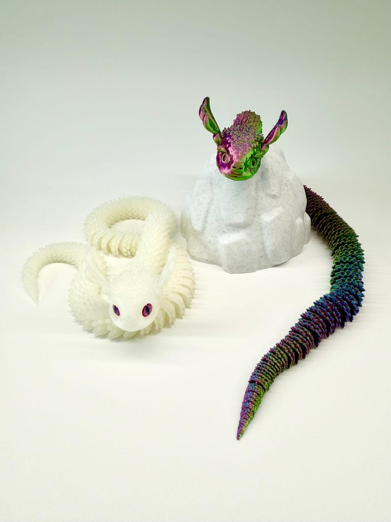 May include: Two fantasy dragon sculptures. One is white with pink eyes, coiled on the surface. The other has a rainbow tail and a head with green, purple, and pink hues, perched on a white rock-like structure.