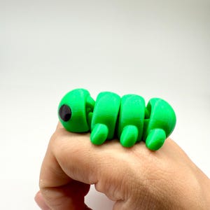 Könnte beinhalten: Ein leuchtend grünes, bewegliches Raupenspielzeug mit einem schwarzen Auge. Das Spielzeug besteht aus Kunststoff und wird vor einem weißen Hintergrund gezeigt. Die Raupe ist segmentiert und scheint 3D-gedruckt zu sein.