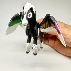 May include: A 3D-printed Pegasus figurine with a black body, white mane, and white wings with iridescent accents. The toy horse has articulated joints and is held against a white background. The wings are decorated with green, blue, and purple hues.