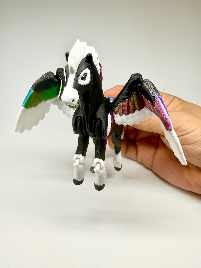 May include: A 3D-printed Pegasus figurine with a black body, white mane, and white wings with iridescent accents. The toy horse has articulated joints and is held against a white background. The wings are decorated with green, blue, and purple hues.