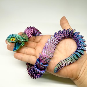May include: A colorful, articulated snake toy with a detailed design. The snake features a vibrant gradient of colors, including green, blue, purple, and pink. The snake's head has yellow eyes and a detailed mouth. The toy is held in a hand.