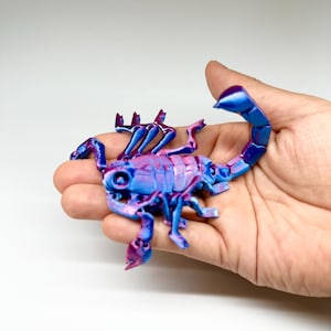 Könnte beinhalten: Eine 3D-gedruckte Skorpionfigur in Blau- und Lilatönen. Der Skorpion wird in einer Hand gehalten und zeigt seinen detaillierten, segmentierten Körper, die Zangen und den gebogenen Schwanz. Die Figur hat eine glänzende Oberfläche und steht vor einem schlichten weißen Hintergrund.