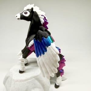 May include: A black and white pegasus figurine with a white mane and wings. The wings have blue, green, and purple accents. The figurine is standing on a textured, white base. The pegasus has a purple gem eye.