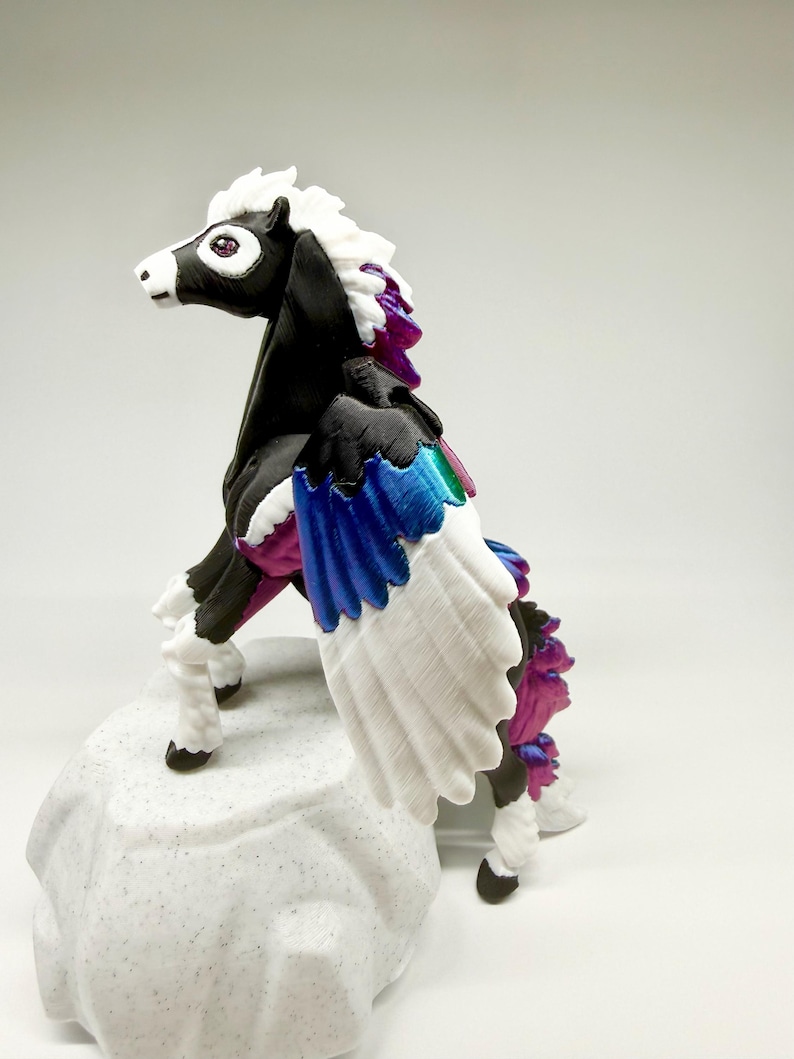 May include: A black and white pegasus figurine with a white mane and wings. The wings have blue, green, and purple accents. The figurine is standing on a textured, white base. The pegasus has a purple gem eye.