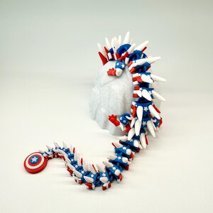 Puede incluir: Un juguete de drag&oacute;n articulado en rojo, blanco y azul, con un escudo y estrellas. El juguete est&aacute; sobre una superficie blanca, destacando su dise&ntilde;o flexible y colores vibrantes. Un dise&ntilde;o patri&oacute;tico y &uacute;nico.