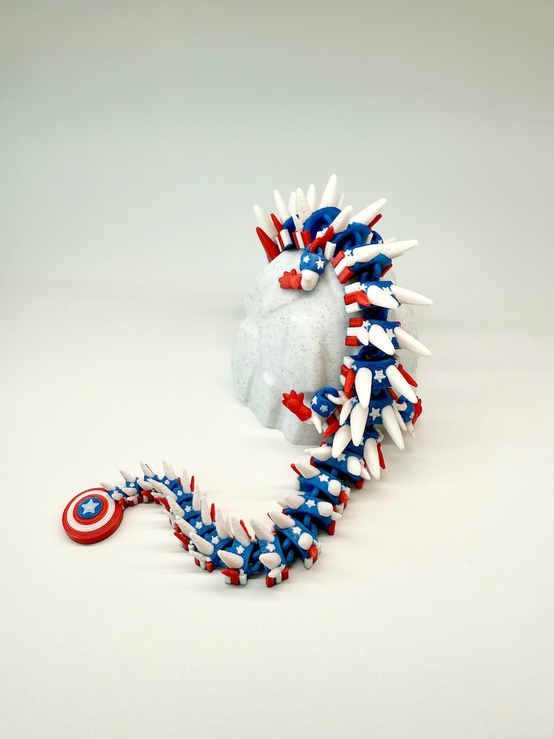 Puede incluir: Un juguete de drag&oacute;n articulado en rojo, blanco y azul, con un escudo y estrellas. El juguete est&aacute; sobre una superficie blanca, destacando su dise&ntilde;o flexible y colores vibrantes. Un dise&ntilde;o patri&oacute;tico y &uacute;nico.