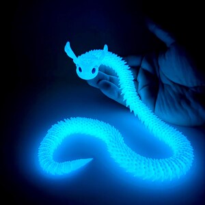 May include: A glowing blue articulated snake toy with a smiling face and small ears. The toy is held in a hand against a dark background. The snake's body is segmented and textured, emitting a bright, cool light.