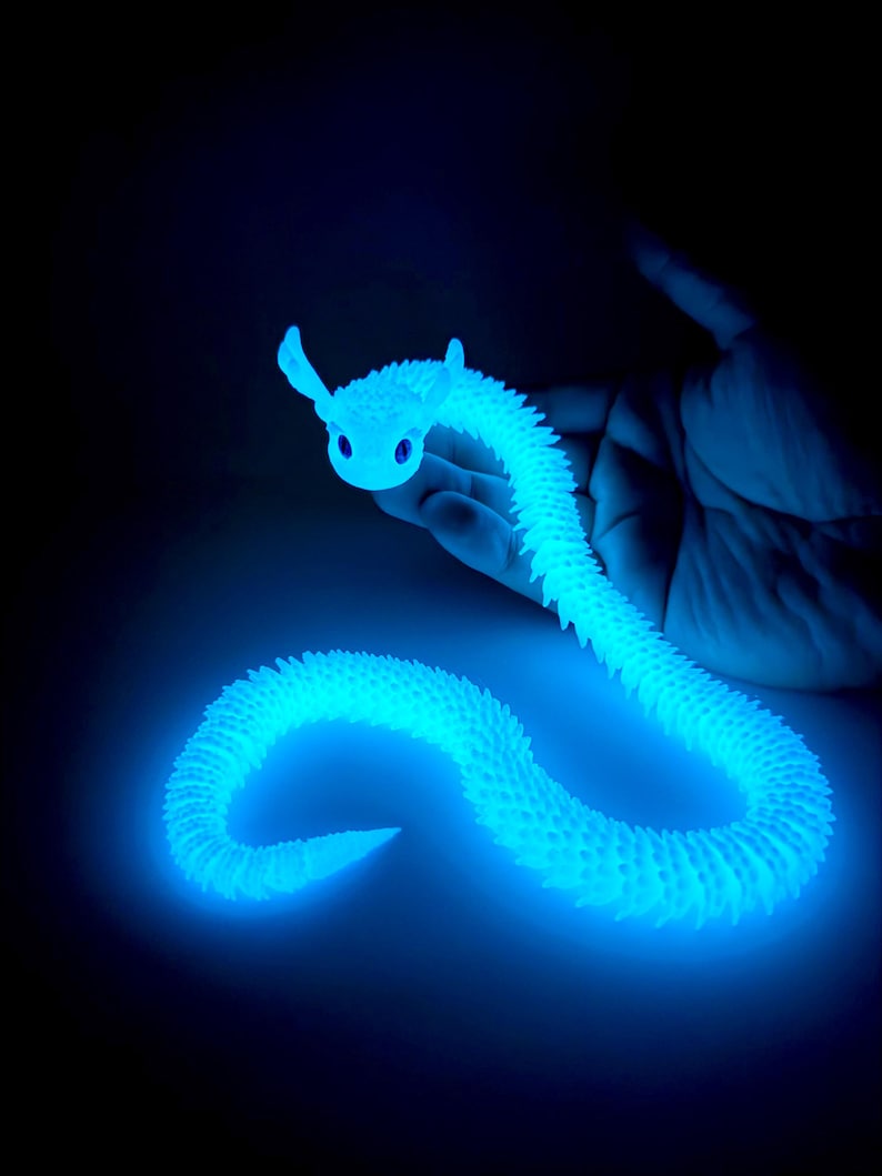 May include: A glowing blue articulated snake toy with a smiling face and small ears. The toy is held in a hand against a dark background. The snake's body is segmented and textured, emitting a bright, cool light.