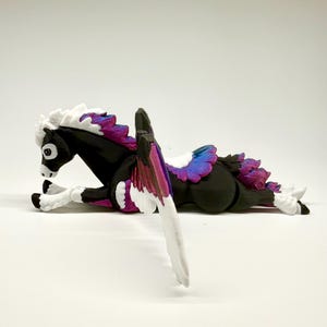 May include: A black Pegasus figurine with white, purple, and pink wings and mane. The mythical creature is lying down, with its wings spread. The figurine has a white chest and legs, and a black body. The Pegasus is a fantasy-themed item.