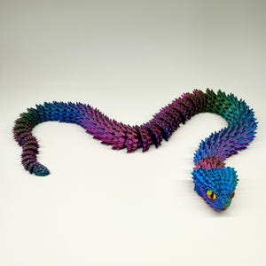 May include: A colorful, articulated snake toy in shades of blue, purple, and green. The snake has a textured, scale-like pattern and a detailed head with yellow eyes. The toy is posed on a white surface.