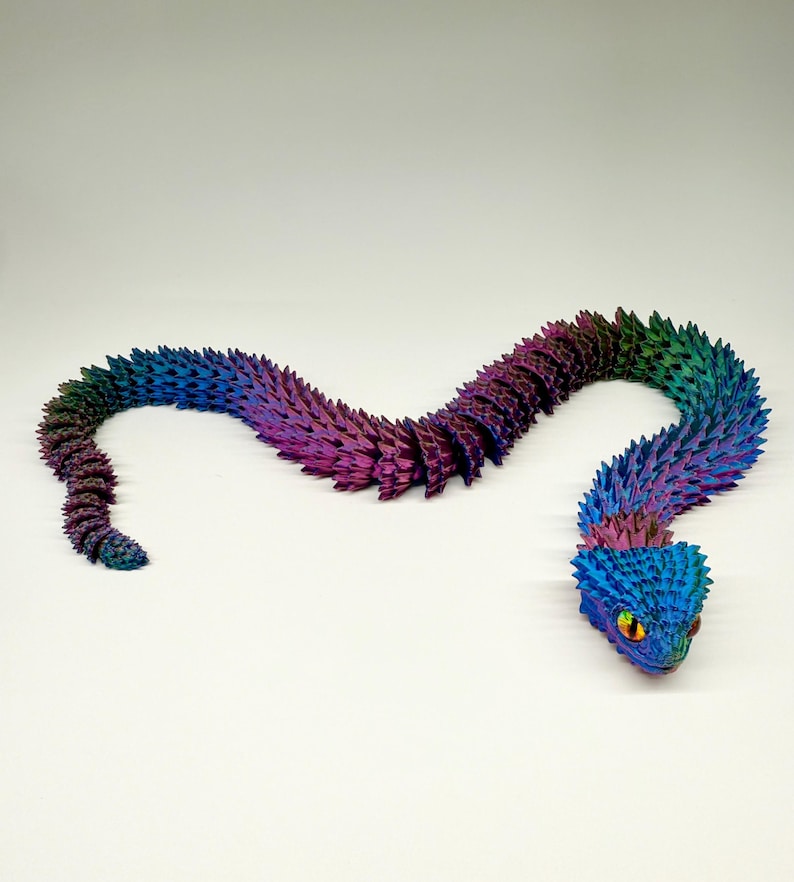 May include: A colorful, articulated snake toy in shades of blue, purple, and green. The snake has a textured, scale-like pattern and a detailed head with yellow eyes. The toy is posed on a white surface.