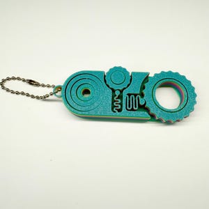 以下が含まれることがあります： 虹色の光沢のある、ターコイズ色のマルチツールキーホルダー。ツールは、円や歯車のようなデザインなど、さまざまな幾何学的な形状を特徴としています。小さな金属製のボールチェーンが付いています。