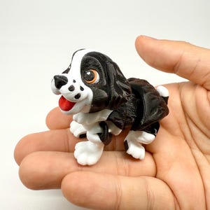 Puede incluir: Una pequeña figura de perro blanco y negro con lengua roja y ojos naranjas. El cachorro tiene patas blancas y un patrón de manchas blancas y negras. La figura se sostiene en una mano abierta.