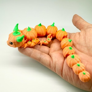 Könnte beinhalten: Ein 3D-gedrucktes orangefarbenes Kürbisraupenspielzeug mit grünen Stielen und Blättern. Das Spielzeug wird in einer Hand vor weißem Hintergrund gehalten. Die Raupe besteht aus mehreren kürbisförmigen Segmenten.