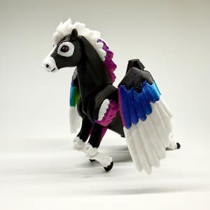 May include: A 3D-printed Pegasus figurine. The black horse has white feathered wings with blue and white tips, and a rainbow gradient on the inner wings. The mane and tail are white and purple. The figurine is on a white background.